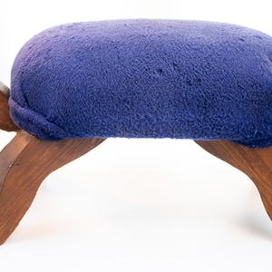 May include: A footstool shaped like a turtle with a dark brown wooden base featuring four legs, a head with a silver eye, and a tail. The top of the turtle is covered in a plush, deep purple fabric cushion that serves as the seat of the stool.