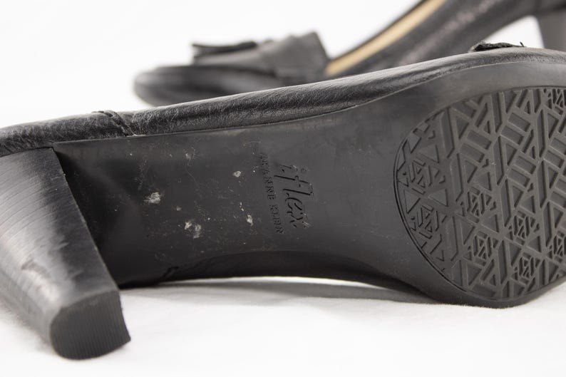 May include: Close-up of a pair of black leather heeled shoes. The sole of the shoe is visible, featuring a textured pattern and the brand name "flex". The heel is thick and sturdy. The other shoe is slightly out of focus in the background.