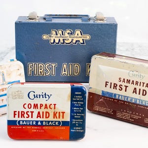 Puede incluir: Un surtido de botiquines de primeros auxilios vintage de varios tamaños y diseños. Los kits incluyen una caja metálica azul con el texto "MSA" y "FIRST AID", un kit "Johnson & Johnson" blanco y azul, y dos kits "Curity", uno rojo y blanco, y otro marrón y blanco.