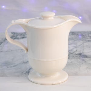 May include: Off-white ceramic pitcher with a lid and handle. The pitcher has a curved spout and a pedestal base. The handle is arched, and the lid has a small knob. The pitcher is likely for serving beverages.