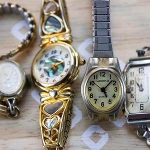 May include: Four wristwatches with various designs. One has a gold-tone band and heart-shaped accents with iridescent inlays. Another is a silver-tone watch with the text watch-it on the face. Two others feature simple oval and rectangular faces.