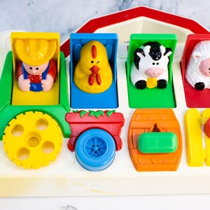 Pode incluir: Um brinquedo pop-up vintage com tema de fazenda. Apresenta um trator verde com um fazendeiro, um pintinho amarelo, uma vaca preta e branca e uma ovelha branca. Inclui rodas amarelas, um telhado de celeiro vermelho e uma cerca amarela.