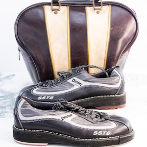 May include: A pair of black and silver Dexter bowling shoes with the text "SST8" and a brown and cream bowling bag. The shoes have black laces and a red accent on the sole. The bag has a brown leather exterior with cream and gold stripes.