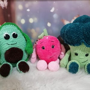 May include: Three crocheted plush toys shaped like an avocado, a strawberry, and a broccoli. The avocado is green with a brown pit and a smiling face. The strawberry is pink with a smiling face. The broccoli is green with a blue head and a smiling face.