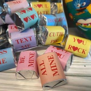 May include: Assortment of small, foil-wrapped chocolate candies with Valentine's Day themed messages printed on the wrappers. The messages include "Kiss Me", "Text Me", "Be Mine", "All Mine", "Cutie Pie", "You Rock", and "Soul Mate".