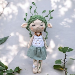 Peut inclure: Poupée au crochet faite à la main avec un design fantaisiste. La poupée a un corps beige, une couronne verte feuillue et une jupe verte. Elle a les yeux fermés, un haut blanc et un collier de perles en bois. La poupée mesure environ 20 cm de haut.
