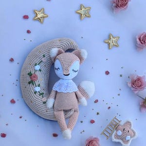Puede incluir: Una muñeca de zorro de ganchillo hecha a mano en rosa suave y blanco, con un cuello y una cola blancos, descansando sobre una luna creciente beige. Acentos de estrellas doradas y rosas rosas rodean la muñeca, creando un tema de guardería caprichoso.
