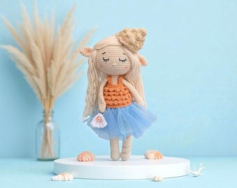 Könnte beinhalten: Handgemachte Häkelpuppe mit langem blondem Haar, die ein orangefarbenes Oberteil, einen blauen Tüllrock und einen muschelförmigen Hut trägt. Die Puppe hält eine kleine weiße Handtasche. Hellblauer Hintergrund, dekorative Muscheln.