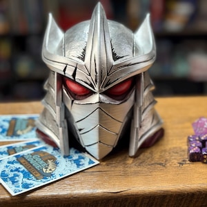 May include: A silver Shredder helmet with red eyes, next to playing cards and purple dice. The helmet has a detailed design with sharp edges. The playing cards have a blue wave pattern and the text "GONE ROGUE: THE GAMES".