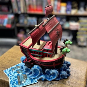 May include: A detailed, red and black model ship with red sails and green dragon figureheads. The ship sits atop blue wave-like structures. Two cards with the text "Gone Rogue Games" are in front of the ship.