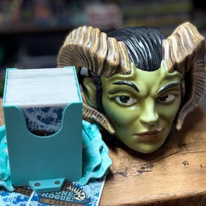 May include: A teal card holder with a sculpted base and a horned demon head. The card holder is filled with white cards and sits on a wooden surface. The demon head is green with brown horns.