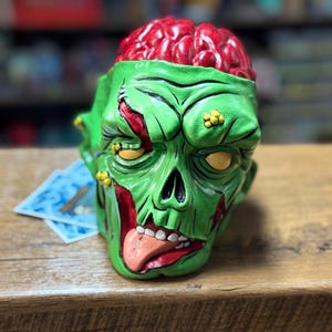 May include: A green zombie head-shaped ceramic container with a red brain exposed at the top. The zombie has yellow eyes, a red mouth with a tongue sticking out, and red gashes. The container is on a wooden surface.