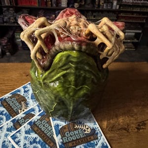 May include: A grotesque, multi-textured sculpture with a green base and a fleshy, bone-like top. Several playing cards with a blue wave design and the text "GONE ROGUE! GAMES" are in the foreground. The sculpture is on a wooden surface.