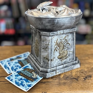 May include: A detailed, gray and gold decorative tabletop fountain with a dragon design. The fountain has a square base and a bowl-shaped top. Three blue and white cards with the text "Gone Rogue! Games" are in front of the fountain.