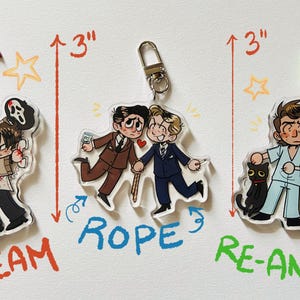 May include: Three acrylic keychains, each approximately 3 inches tall, featuring cartoon illustrations. The keychains depict characters from horror films, including "Scream," "Rope," and "Re-Animator." Each keychain has a different colored clasp.
