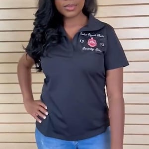 Puede incluir: Una camiseta polo negra con un logotipo bordado en blanco que dice "Delta Sigma Theta Sorority, Inc." con los números "19" y "13" a cada lado del logotipo. La camiseta la lleva puesta una persona que lleva pantalones vaqueros azules y gafas de sol negras.