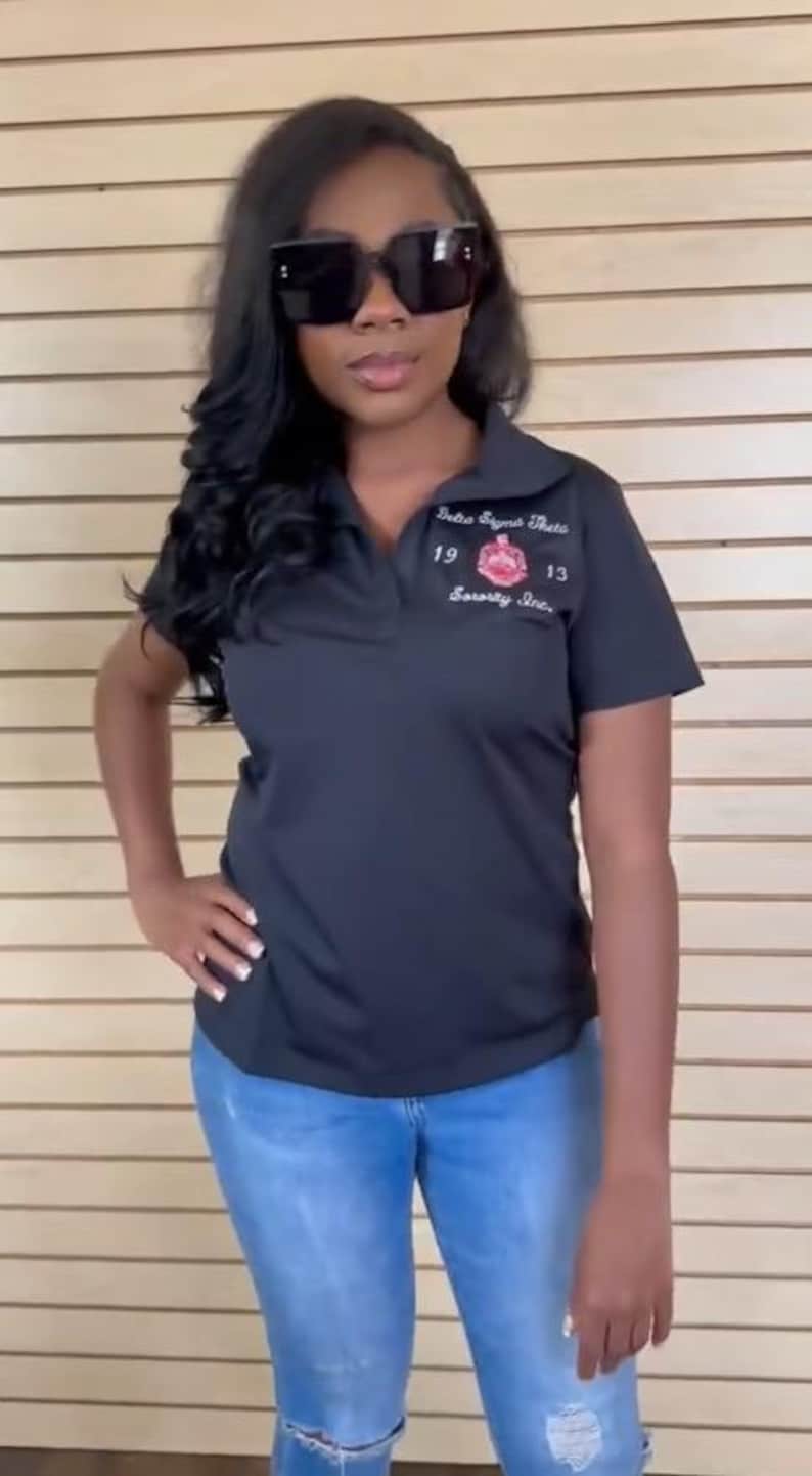 May include: A black polo shirt with a white embroidered logo that says "Delta Sigma Theta Sorority, Inc." with the numbers "19" and "13" on either side of the logo. The shirt is being worn by a person wearing blue jeans and black sunglasses.