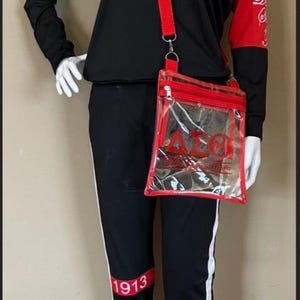 May include: Black and white tracksuit with red accents. The top has the letters "AEO" in red on the chest. The pants have a red stripe with the number "1913" on the left leg. A clear plastic bag with a red zipper and red trim has the letters "AEO" in red on the front. The bag is attached to a red strap.