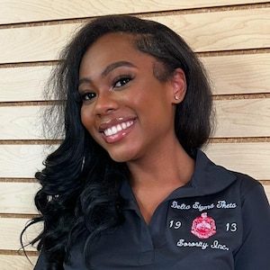 May include: A woman with long black hair is smiling and wearing a black polo shirt with the text "Delta Sigma Theta Sorority Inc." and the numbers "19" and "13" embroidered on it.
