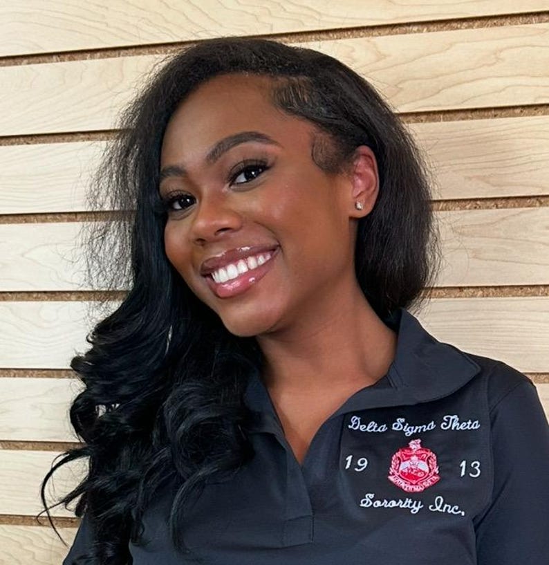 May include: A woman with long black hair is smiling and wearing a black polo shirt with the text "Delta Sigma Theta Sorority Inc." and the numbers "19" and "13" embroidered on it.