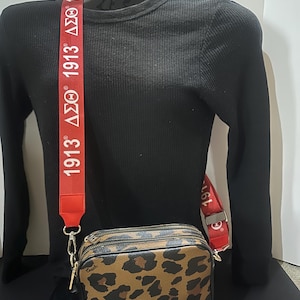 May include: A brown and black leopard print Coach crossbody bag with a red strap that says "1913 ΔΣΘ 1913 ΔΣΘ".
