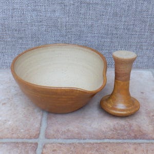 Pode incluir: Um conjunto de almofariz e pilão em cerâmica, composto por uma taça com interior creme salpicado e exterior castanho-dourado. O pilão apresenta um acabamento a condizer em castanho-dourado, com uma base alargada e um cabo cilíndrico.
