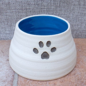 Puede incluir: Tazón de cerámica blanco para mascotas con interior azul y diseño de huella de pata gris.