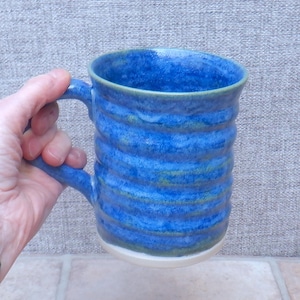 Op de afbeelding: Handgemaakte blauwe keramische mok met een getextureerd, geribbeld ontwerp. De mok heeft een groot handvat en een witte basis. De binnenkant en de rand hebben een donkerdere blauwe tint, met hints van groen. Een uniek, handgemaakt stuk.
