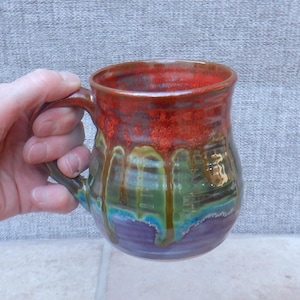 May include: A handmade ceramic mug with a unique glaze. The top of the mug is a vibrant red, transitioning to green and blue, and ending with a purple base. The mug has a sturdy handle and a glossy finish.