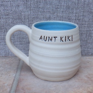 Puede incluir: Taza de cerámica blanca con interior azul y las palabras "AUNT KIKI" escritas en el lateral.