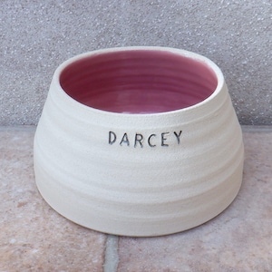 Puede incluir: Tazón de cerámica blanco para mascotas con interior rosa y el nombre "Darcey" grabado en la parte delantera.