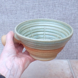 Puede incluir: Un cuenco de afeitado de cerámica hecho a mano con un asa. El cuenco tiene un interior azul claro con un patrón de rejilla y un exterior marrón y marrón. El cuenco se sostiene en una mano.