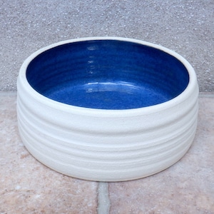 Puede incluir: Tazón de cerámica blanco con interior esmaltado en azul. El tazón tiene una forma redondeada y un exterior ligeramente texturizado.