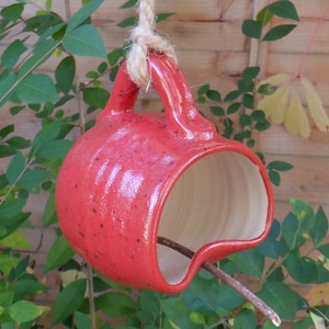 May include: A red ceramic bird feeder shaped like a mug with a hole cut out for birds to access the food. The feeder is hanging from a rope.