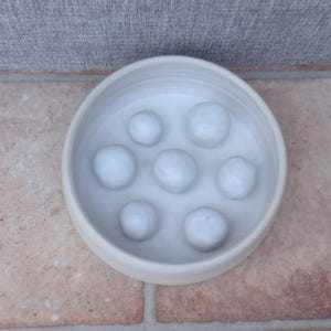 Puede incluir: Un cuenco de cer&aacute;mica de color blanco roto, de forma redonda, con siete protuberancias elevadas, lisas y redondeadas de distintos tama&ntilde;os en la base interior. El cuenco presenta un dise&ntilde;o sencillo con un acabado mate y un color crema uniforme.