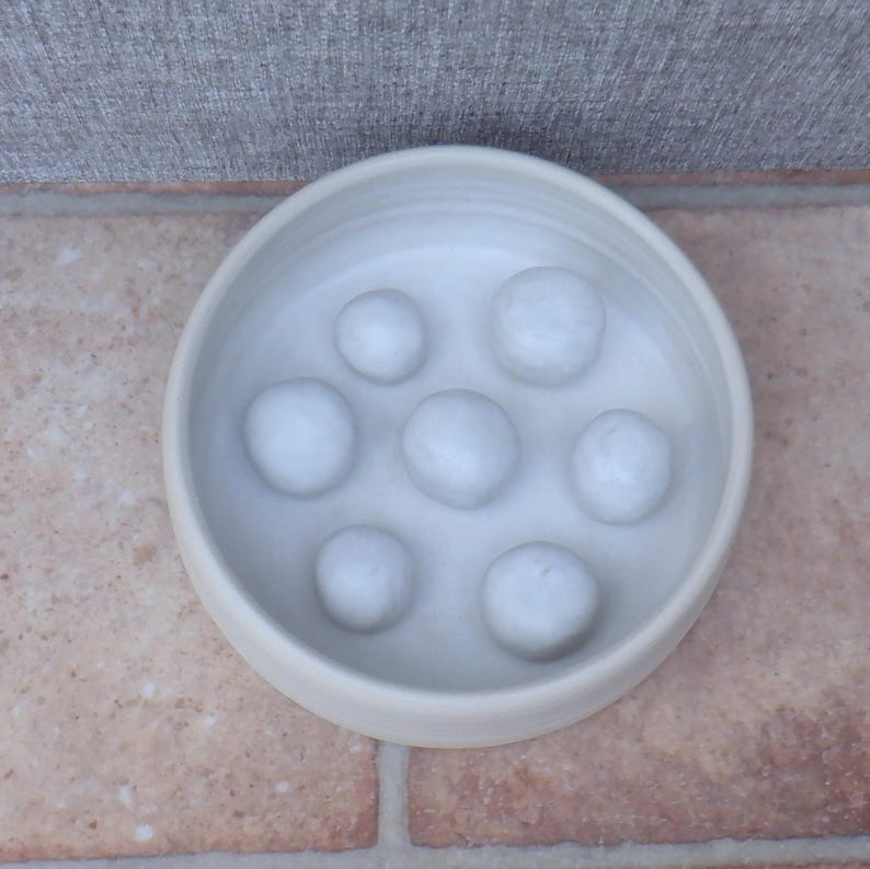 Puede incluir: Un cuenco de cer&aacute;mica de color blanco roto, de forma redonda, con siete protuberancias elevadas, lisas y redondeadas de distintos tama&ntilde;os en la base interior. El cuenco presenta un dise&ntilde;o sencillo con un acabado mate y un color crema uniforme.