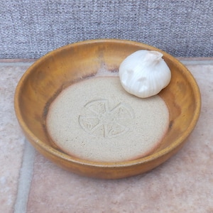 May include: A yellow ceramic bowl with a white glaze and a raised design in the center. A single garlic clove sits in the bowl.
