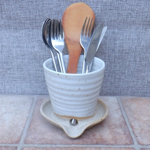 May include: A white ceramic utensil holder with horizontal ridges and a speckled finish, resting on a matching circular base with a small drainage spout. The holder contains several stainless steel forks, a knife, and a wooden spoon.