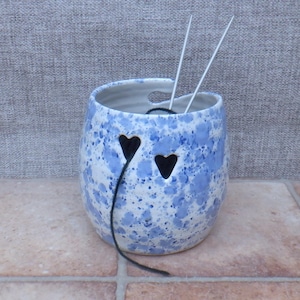 May include: A ceramic yarn bowl with a white base and blue speckled pattern. The bowl features two heart-shaped cutouts on the front and a curved opening at the top rim. Two silver-toned knitting needles are inserted into the bowl, with a single strand of black yarn threaded through one of the heart cutouts.