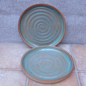 Puede incluir: Dos platos de cerámica hechos a mano. Los platos tienen una combinación de colores turquesa y marrón con un diseño en espiral en el centro. Los bordes son de color marrón oscuro. Los platos están sobre una superficie embaldosada.