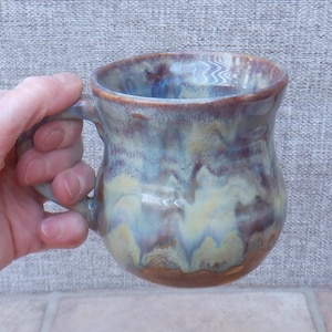 Op de afbeelding: Een handgemaakte keramische mok met een afgeronde vorm en een stevig handvat. De mok heeft een mix van kleuren, waaronder bruin, blauw en crème, waardoor een gemarmerd effect ontstaat. De mok wordt door een hand vastgehouden.