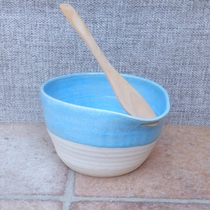 Könnte beinhalten: Eine Rührschüssel aus Keramik mit einem leuchtend blau glasierten oberen Teil und einem strukturierten, cremeweißen unglasierten Boden. Die Schüssel hat einen kleinen Ausgießer am Rand und enthält einen glatten, hellen Holzlöffel im Inneren.