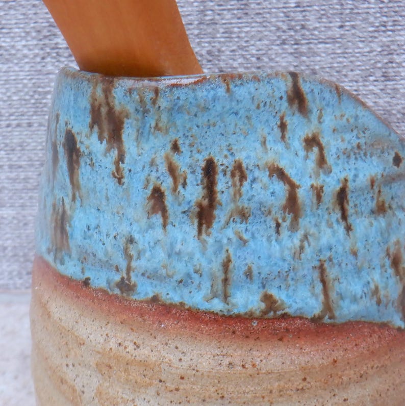May include: A handcrafted ceramic mug with a distinctive design. The mug showcases a light blue glaze with brown speckles and a reddish-brown band near the base. The lower part of the mug has a natural, unglazed finish.