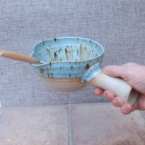 Puede incluir: Cuenco de cerámica hecho a mano con interior azul claro y exterior beige. El cuenco tiene un asa y una cuchara de madera. El cuenco está decorado con motas marrones. El asa la sostiene una persona.