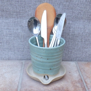 May include: A ceramic utensil holder in a light teal color, holding silverware and a wooden spoon. The holder sits on a matching ceramic spoon rest. The holder and spoon rest have a speckled finish.