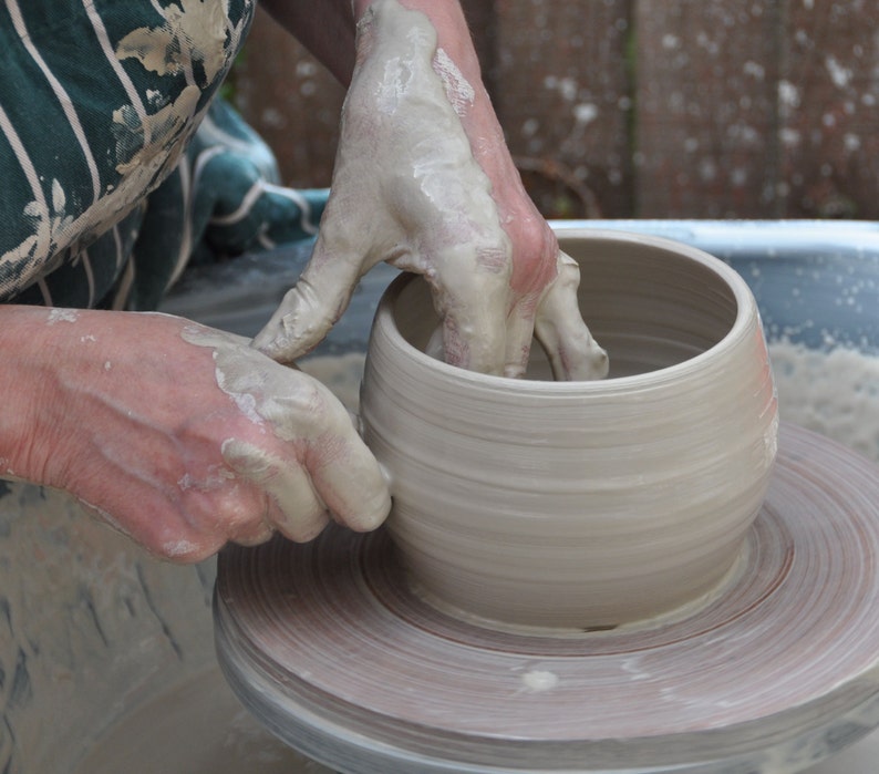 May include: A light grey clay vessel being shaped on a pottery wheel. The circular object features smooth, horizontal ridges along its exterior walls and a wide, open top rim, showcasing the raw, wet texture of unglazed ceramic material.