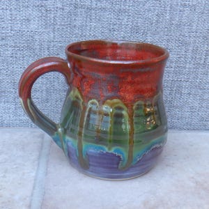 May include: A handmade ceramic mug with a vibrant, multi-coloured glaze. The top is a deep red, transitioning to green, blue, and purple. The handle is a matching red-brown. The mug is approximately 10 cm tall.