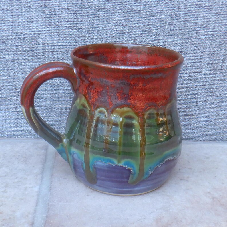 May include: A handmade ceramic mug with a vibrant, multi-coloured glaze. The top is a deep red, transitioning to green, blue, and purple. The handle is a matching red-brown. The mug is approximately 10 cm tall.