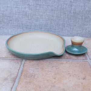 Könnte beinhalten: Handgefertigte Keramik-Löffelablage und passender Deckel. Die Löffelablage ist beige gesprenkelt mit einem türkisfarbenen Rand und einem rotbraunen Akzent. Der Deckel ist klein und rund, mit türkisfarbenem Boden und beigem Oberteil.