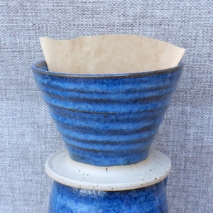 Könnte beinhalten: Handgefertigter blauer Keramik-Kaffee-Pour-Over mit Papierfilter. Der Dripper hat horizontale Rillen und steht auf einem weißen Keramiksockel. Das Design ist tiefblau mit einer rustikalen, handwerklichen Ästhetik.